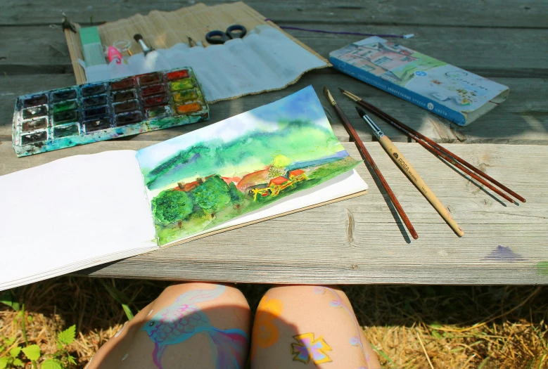 picture sitting on table with supplies including art supplies, brush, and book