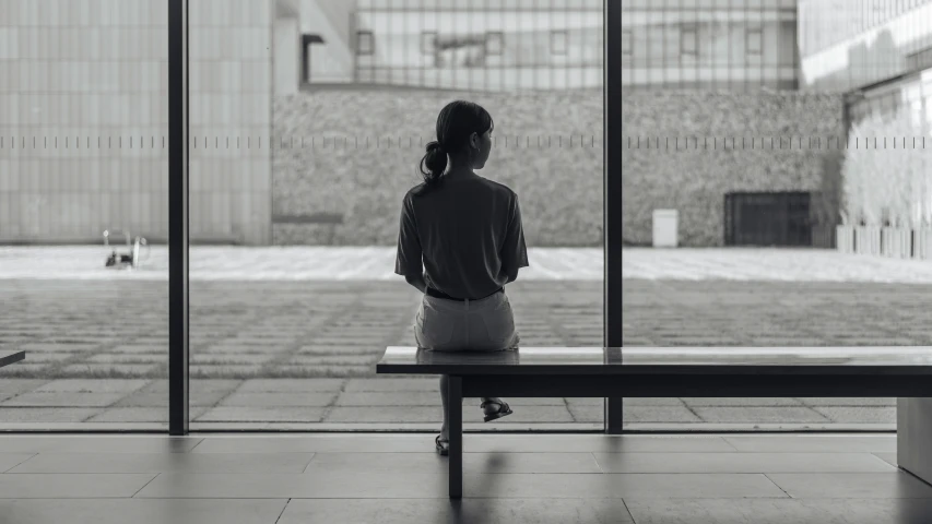 the woman sits on the bench looking out the large glass door