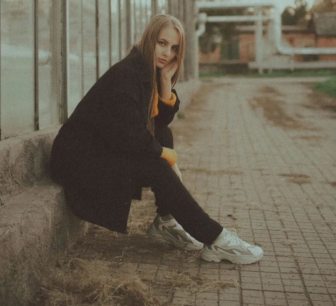 young woman sitting on side of building next to dirt walkway