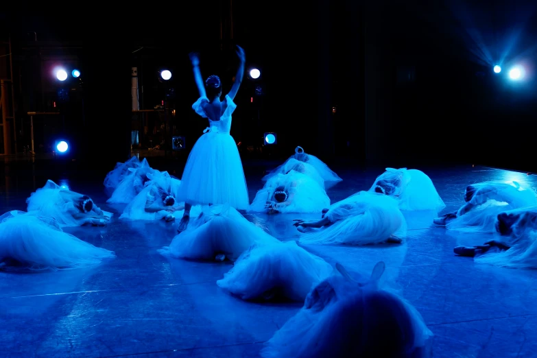 a woman in a white dress stands on stage surrounded by other women and has her arms in the air