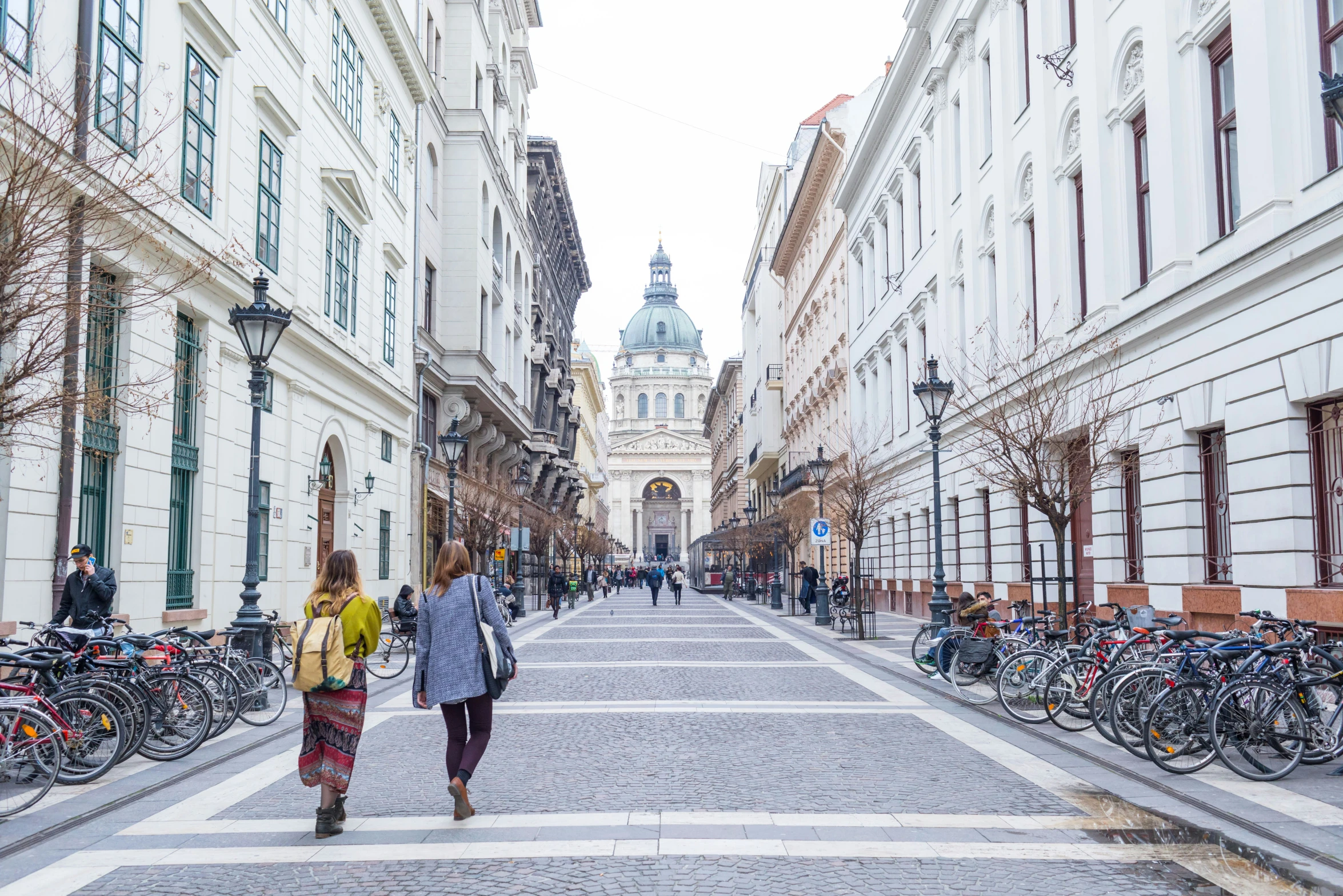 two women walk down a street in front of tall white buildings