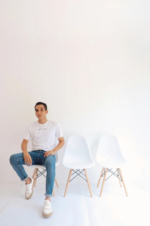 a man sitting in four white chairs with white walls behind him