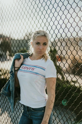 a beautiful young blonde woman in a white shirt and jeans leaning against a fence