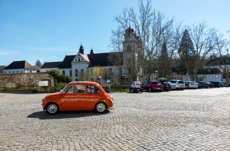 an orange car sits on the cobblestones in front of buildings