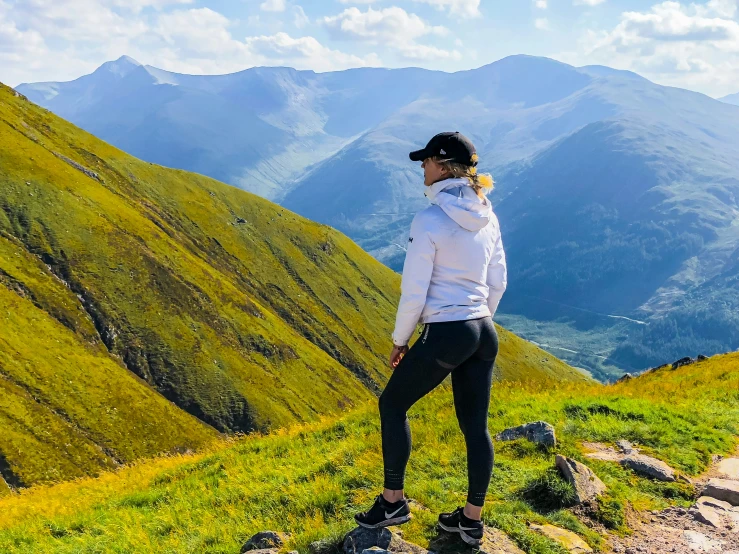 a woman in white jacket and black pants standing on hill with mountains