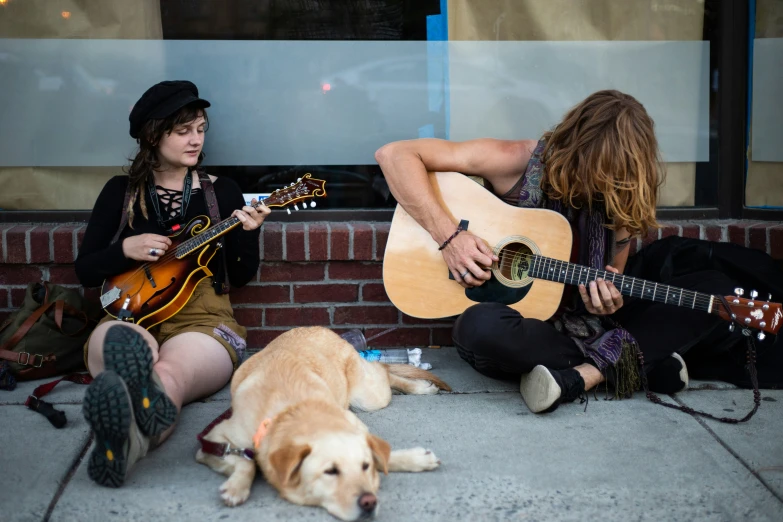 two women sitting on a sidewalk playing guitar, one in front of a dog