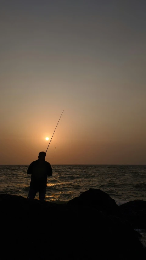 silhouette of person standing on rock with fishing pole at dusk