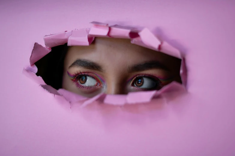 a woman with blue eyes peers out from behind a pink paper