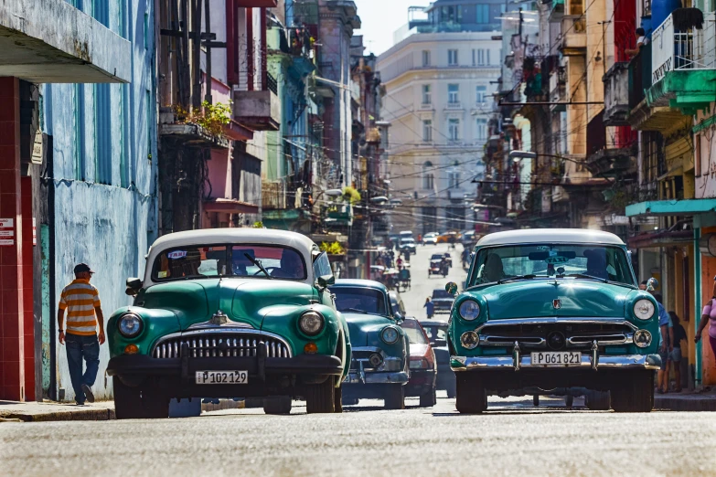 two classic cars drive through an alleyway with a shop on one side and some buildings on the other