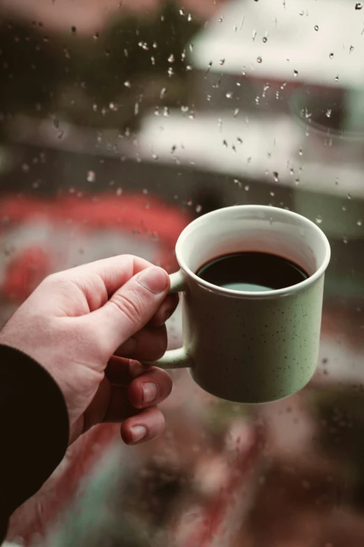 a hand holding a cup of coffee over a rainy window