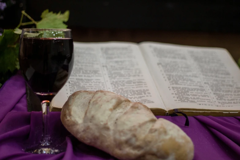 a piece of bread sits next to a glass of wine and an open book