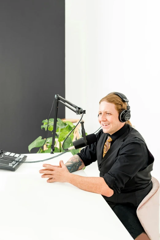 a smiling man with headphones sitting at a white desk with a microphone