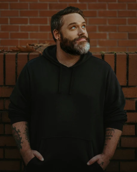 a bearded man wearing a black hoodie standing in front of a brick wall