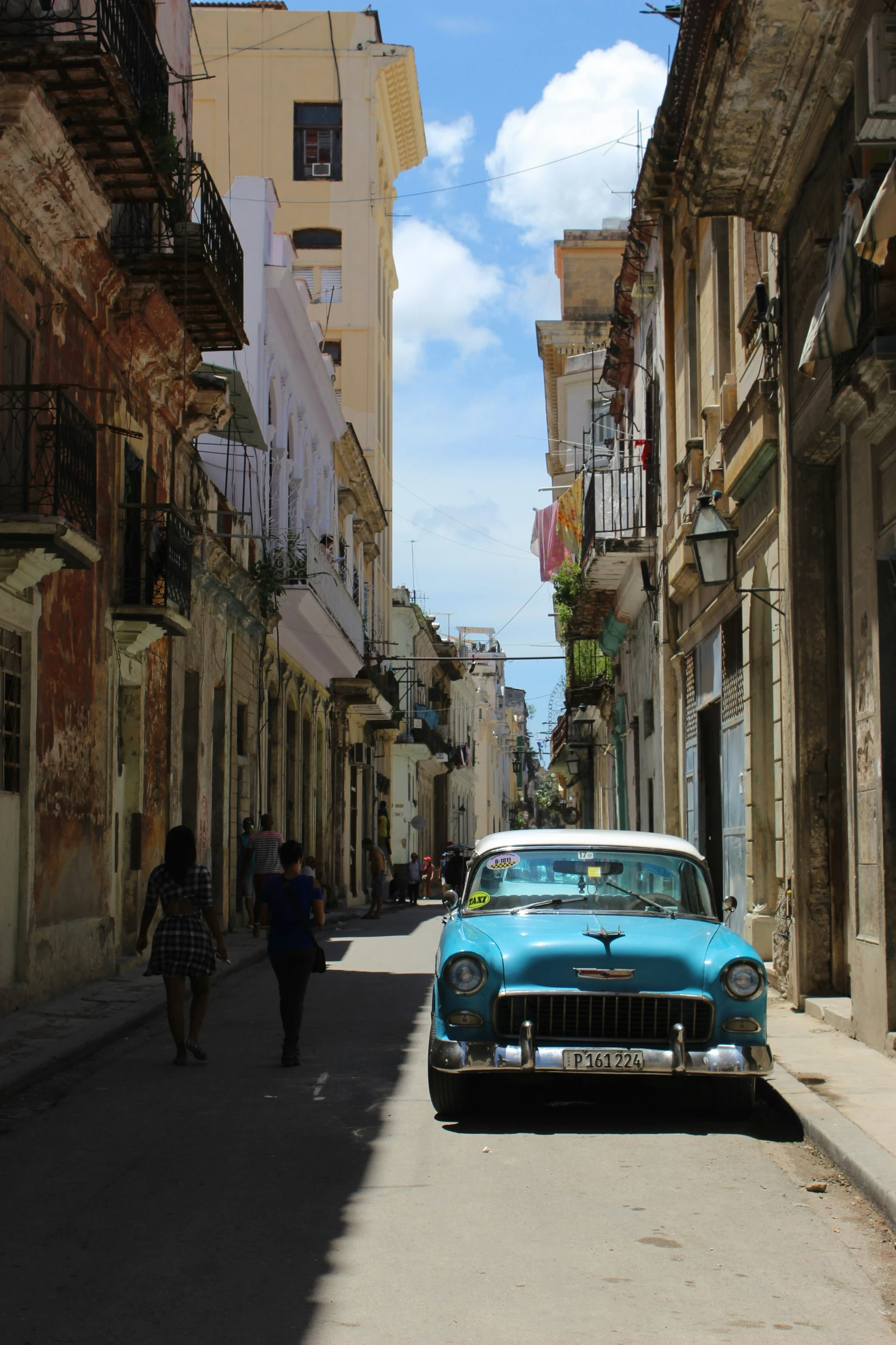 people walk down a small city street in front of an old style car