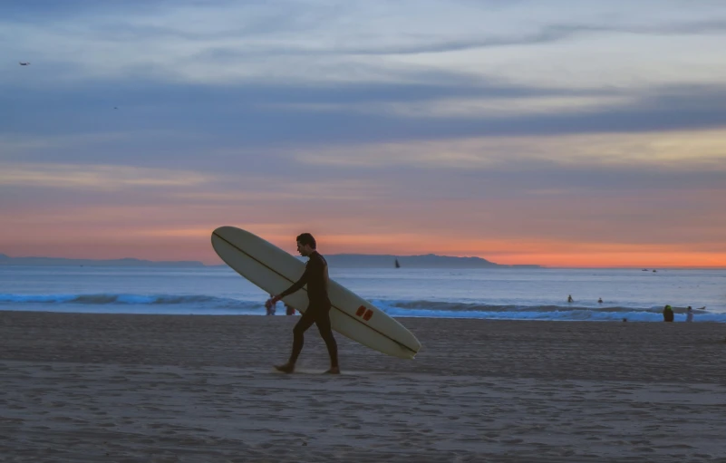 a man walking on a beach carrying a surfboard at sunset