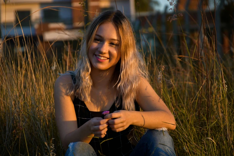 young woman sitting in tall grass in front of some buildings