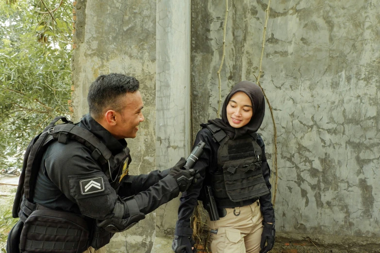 two people, one with black jacket and beige pants, stand near a wall as another person in military gear holds onto the arm of another man