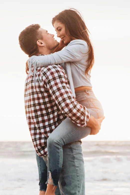 a man in plaid shirt giving a woman piggy back on beach