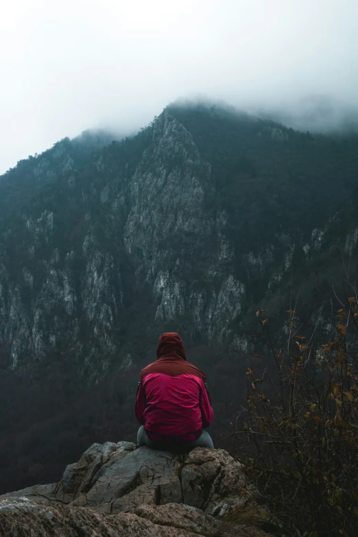 the person is wearing red jacket and sits on the rock near the mountain