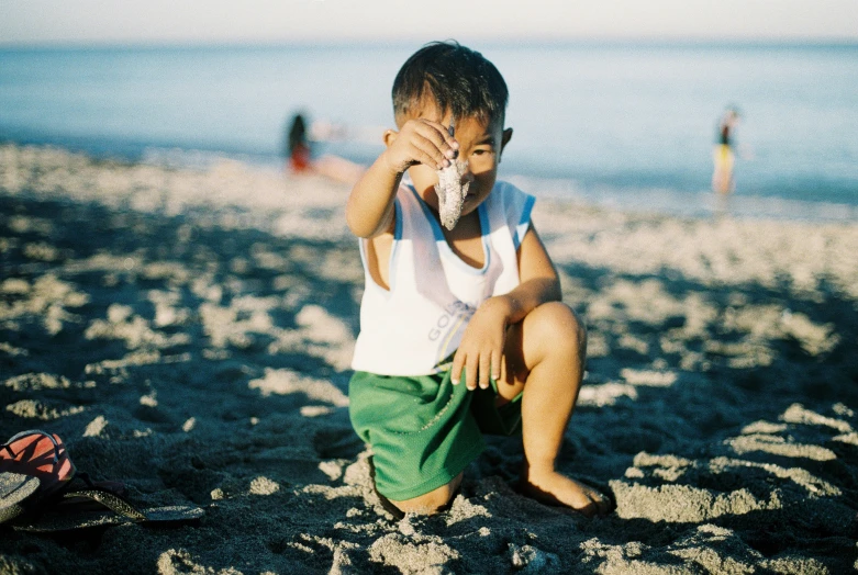 a boy sitting in the sand on the beach with his hand on his face