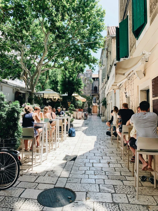 people sit on tables in the shade of trees in an alleyway