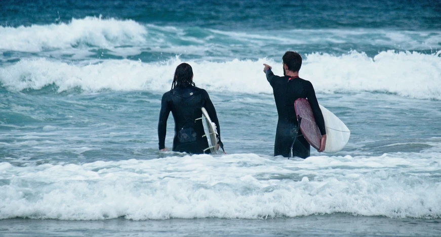 two surfers look out towards the ocean as they walk to shore