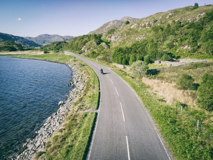 an overhead view of a single motorcycle rider driving on a paved road along the edge of a lake