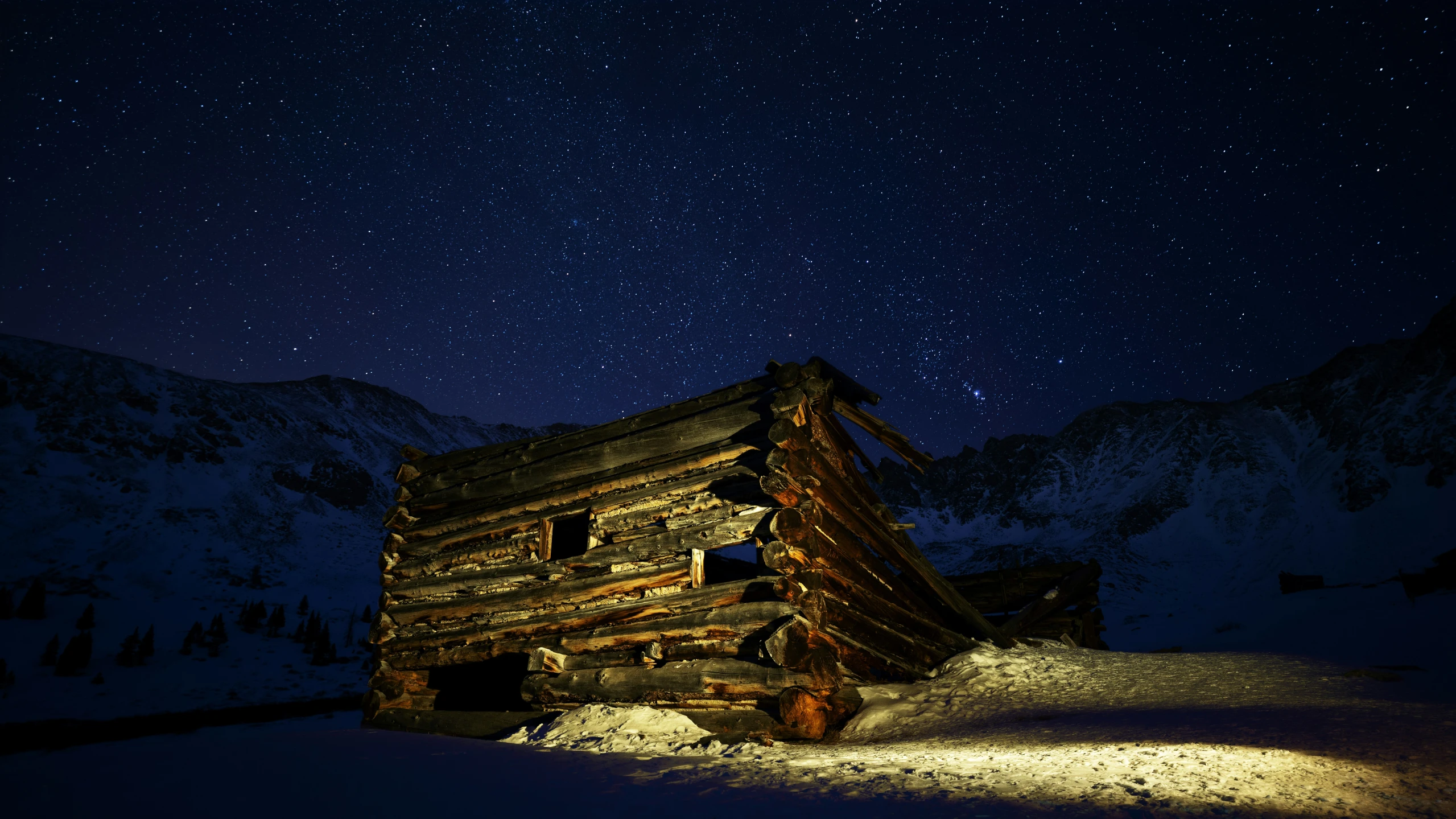 a wooden cabin with snow on the ground below a night sky