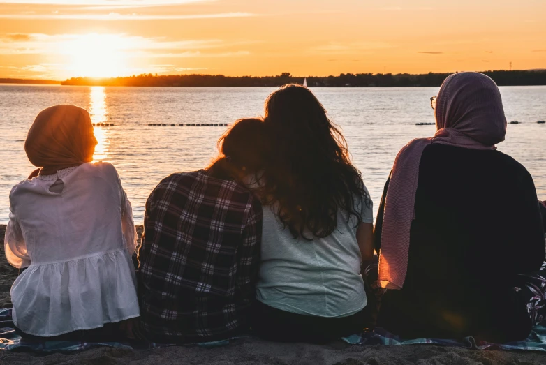 three girls sitting together by the water watching a sunset