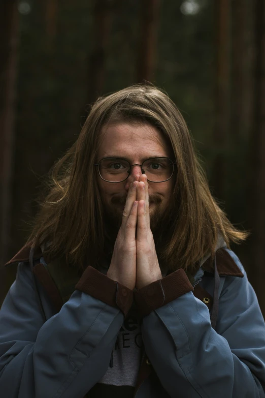 a young man in a blue jacket standing in front of trees holding his hands up to the side of his face