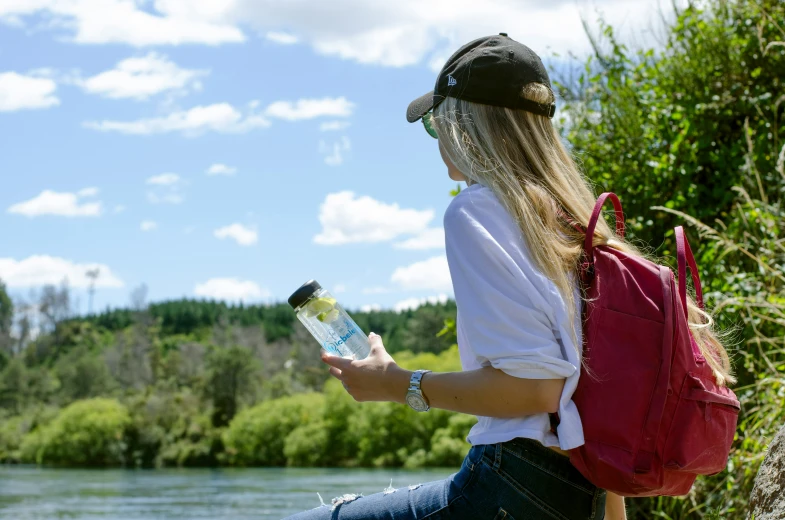 a girl wearing a red backpack holding a water bottle and looking out over the water