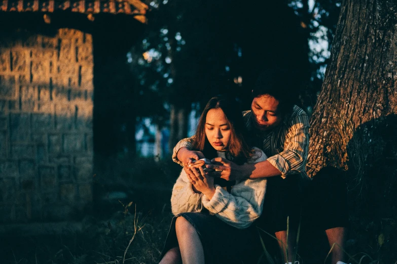 two people sitting under a tree, one of the woman has her hands around the man