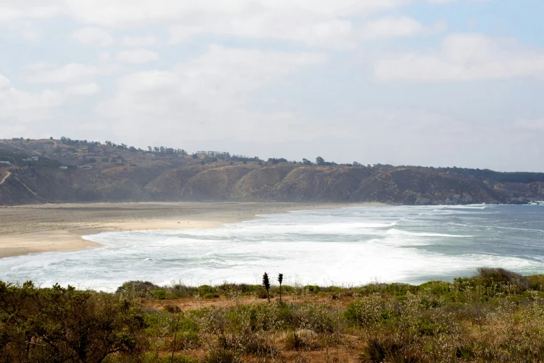 three people stand on a hill above the ocean looking at the beach