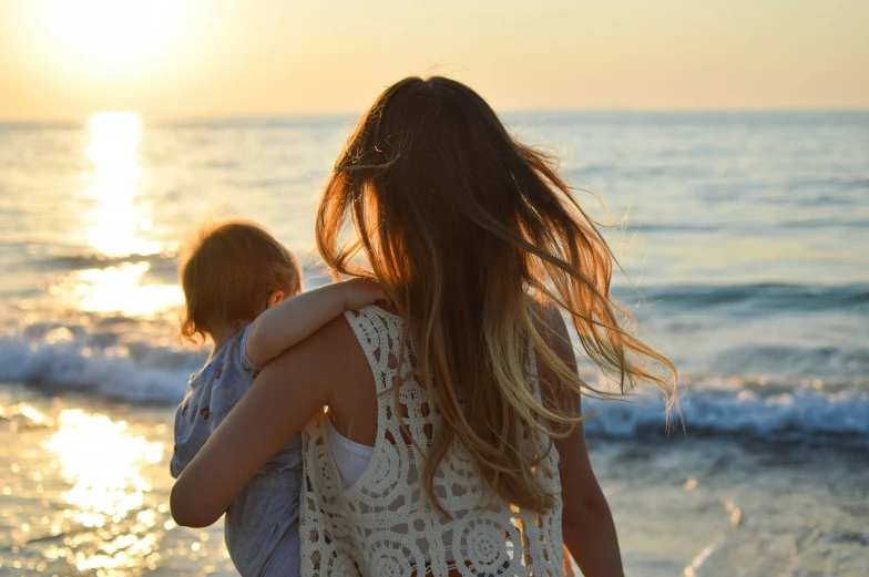 a woman holds a child on the beach as the sun sets