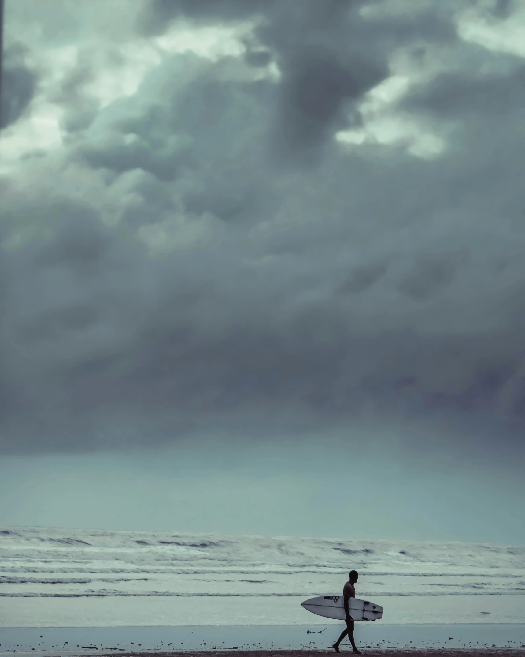 man walking on beach carrying his surfboard with stormy skies above