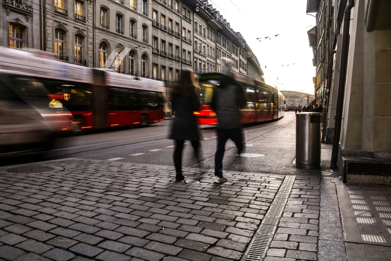 the two people are walking down the cobblestone sidewalk in front of some buildings