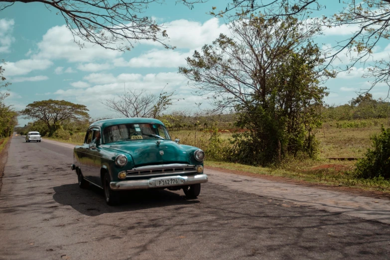 an old green car sitting next to a rural road