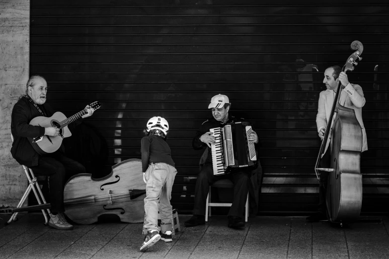 three men and a child sit playing instruments on a city sidewalk