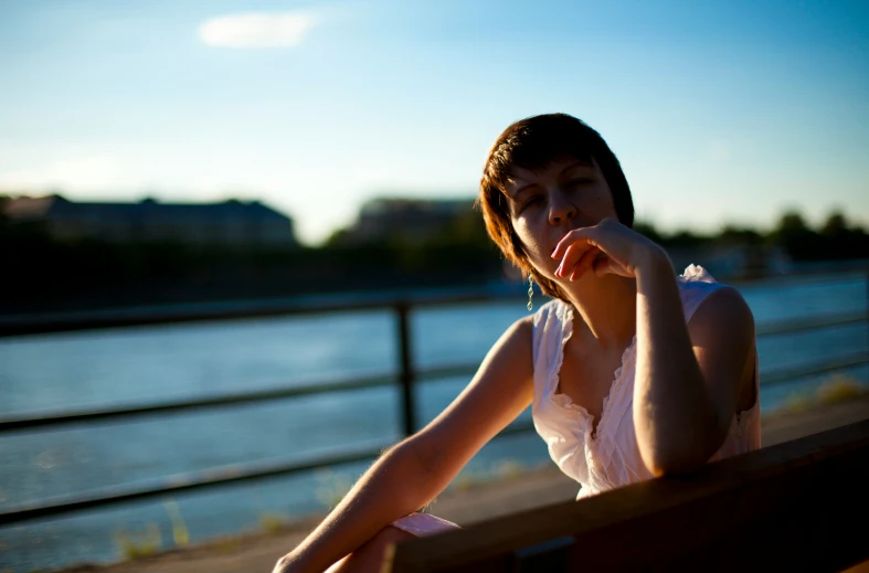 a beautiful young woman sitting on a bench next to water