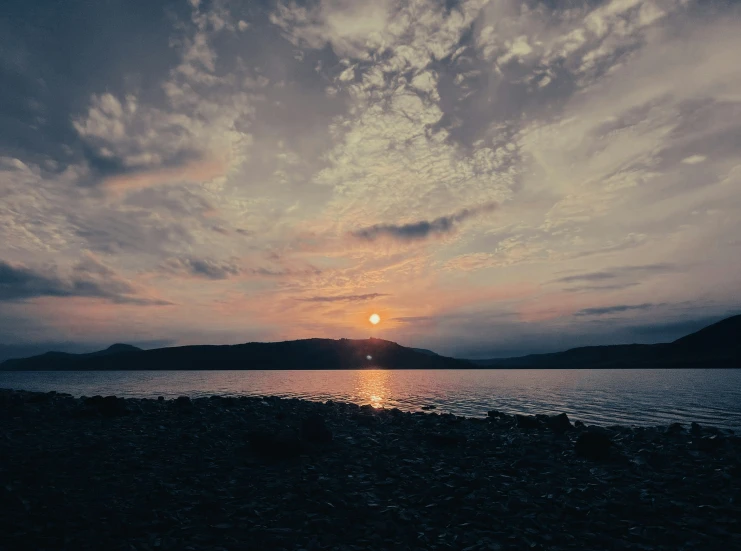 a view of the ocean from a rocky shore with the sun setting over a mountain range