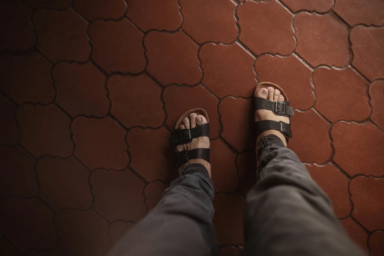 a persons feet wearing black sandals in front of a tiled wall