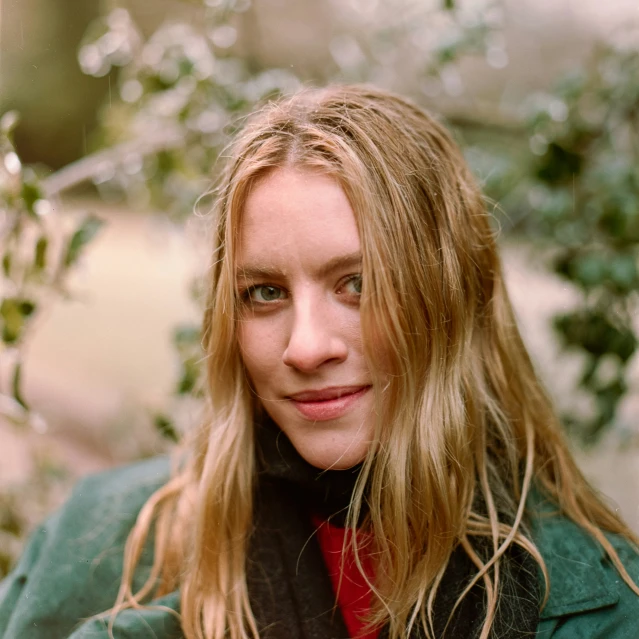 a blonde woman with long hair and green jacket standing next to trees