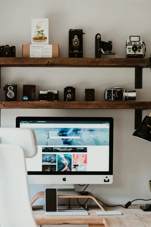 several old cameras are stacked on a wall above a desk with a laptop, monitor and chair