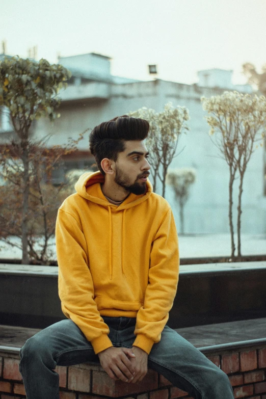 a man with a yellow hoodie and jeans sitting on a brick wall