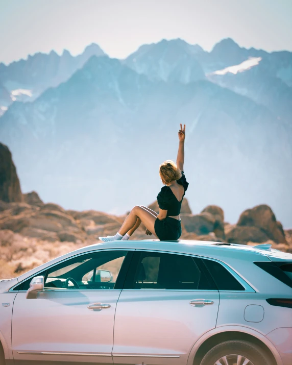 a lady is sitting on the hood of her car and is raising her hands up