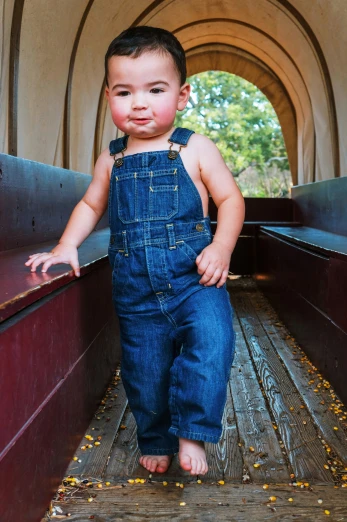 small child in overalls standing on the ground next to a piece of wood