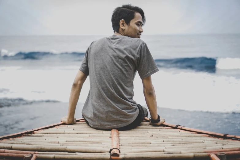 man in grey shirt sitting on bamboo raft with ocean in the background