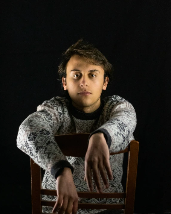 a young man sits down at a table with his hand on the table and finger on the chair