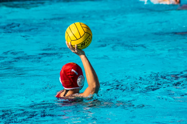 a water polo player in a red cap holds the ball above his head