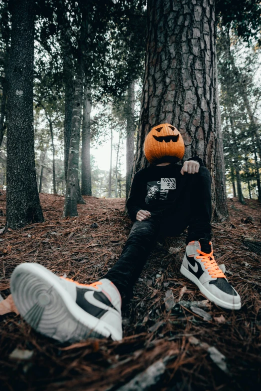a boy sitting in the woods wearing a pumpkin head